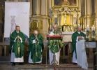 Czterech księży stoi na ołtarzu. Każdy z księży jest ubrany w zielone szaty liturgiczne. Za nimi po lewej widać biały baner. W tle jest złoty ołtarz. 