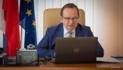 Mężczyzna w garniturze i okularach siedzący za biurkiem na tle okna mówi w kierunku ekranu laptopa. Za nim, po lewej stronie, flaga Polski i Unii Europejskiej.