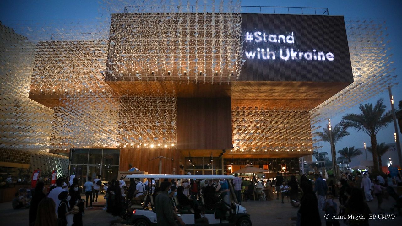 Budynek, bardzo dobrze oświetlony. Na nim ekran led i napisz Stand with Ukraine.