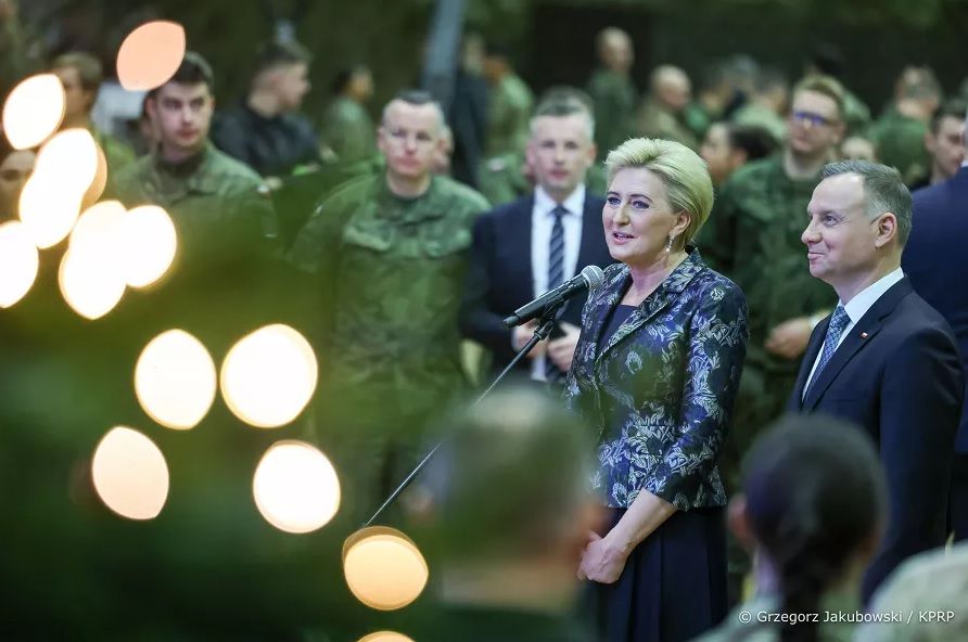 Para prezydencka w otoczeniu żołnierzy. Pierwsza dama mówi do mikrofonu. Na pierwszym planie oświetlona choinka.