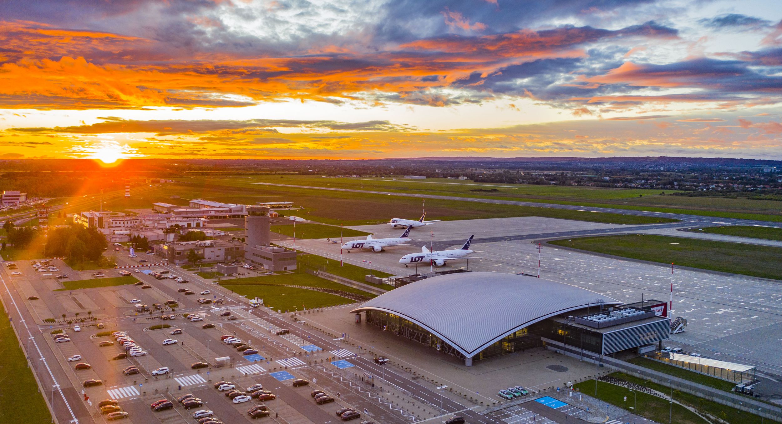 Zdjęcie z lotu ptaka. Na dole Lotnisko: terminal oraz samoloty na płycie lotniczej. W tle Pomarańczowy zachód słońca